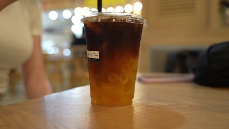 a woman stirring an iced coffee in a plastic cup with a black straw in a cafe, public environment