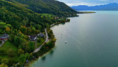 Serena-Orilla-Del-Lago-Con-Veleros,-Rodeada-De-Exuberante-Vegetación-Y-Casas,-Bajo-Un-Cielo-Nublado,-Vista-Aérea