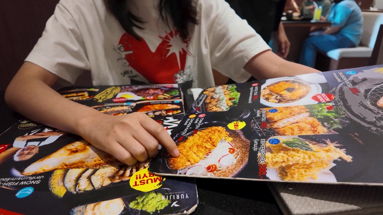 En kvinne peker på tonkatsu-retter på en fargerik meny mens hun spiser i Bangkok, Thailand