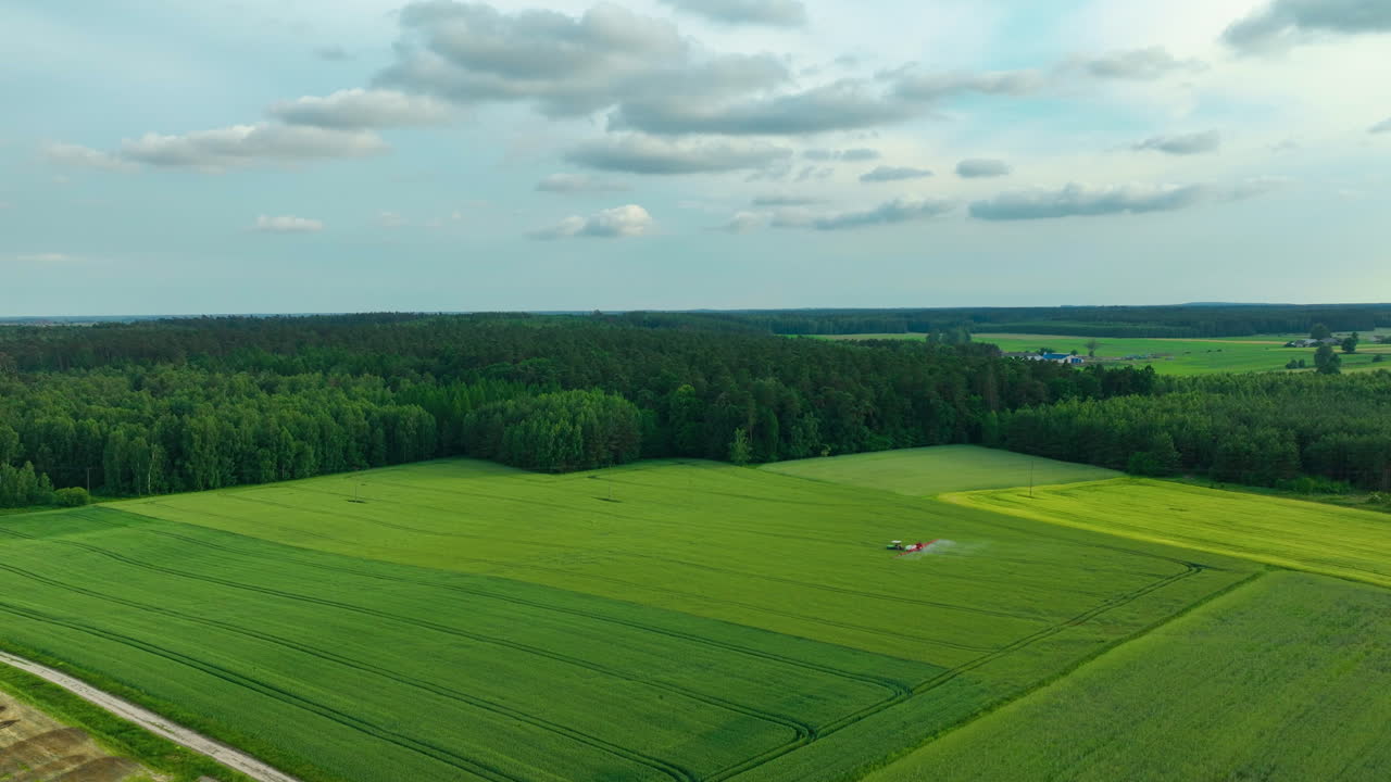 Aerial view of green fields with a tractor working near a forest under a partly cloudy sky