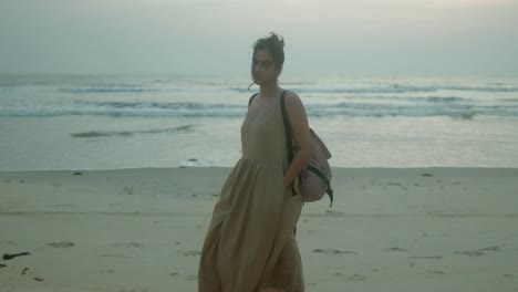 A-woman-in-a-flowing-light-dress,-with-a-backpack,-poses-confidently-on-a-serene-beach-at-sunset