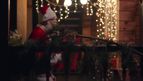 Side-view-Santa-Claus-carries-and-delivers-a-gift-in-his-hands-in-winter-on-the-street-and-comes-to-the-door-and-enters-the-house-with-garlands-and-Christmas-decorations.-In-December-on-Christmas-Eve