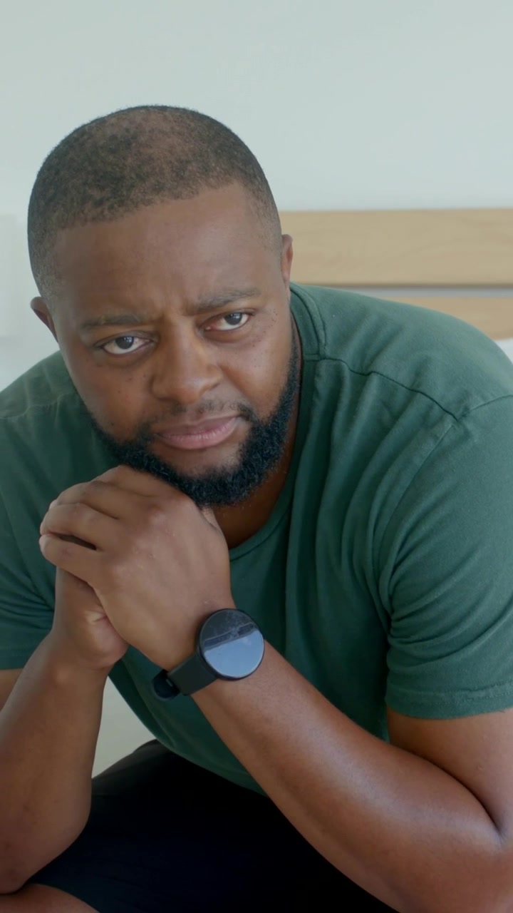 Vertical video: Hearing off-camera voice, man in green T-shirt checking watch while sitting on bed