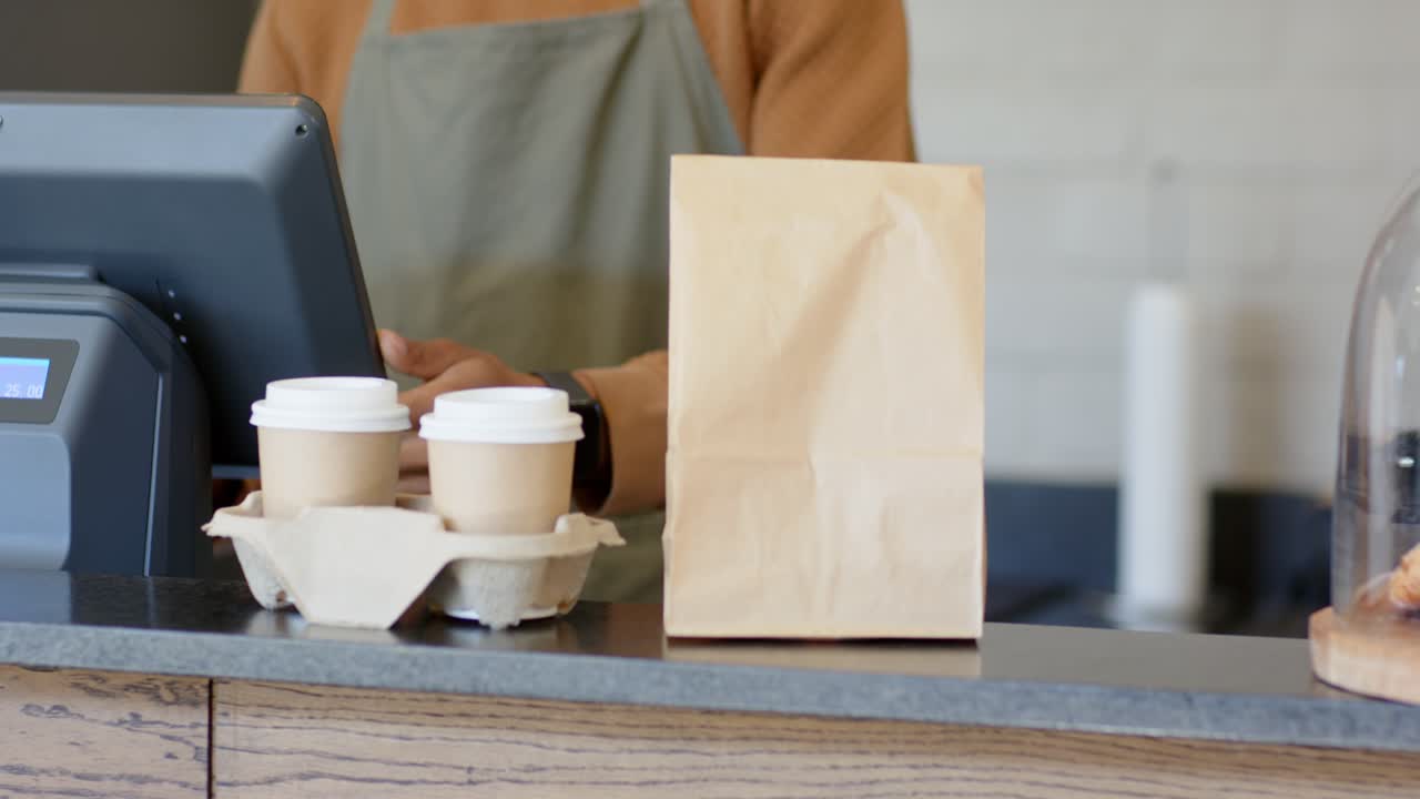 Confirmando o pagamento, barista afro-americano toca na tela, colocando xícaras e uma sacola no balcão de café.