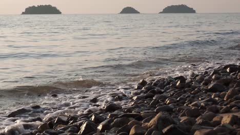 Kleine-Wellen-In-Zeitlupe-Schlagen-Am-Felsigen-Strand-Bei-Sonnenuntergang-Mit-Inseln-In-Der-Ferne