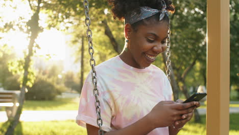 joyous african american woman sitting on swing in park and using phone