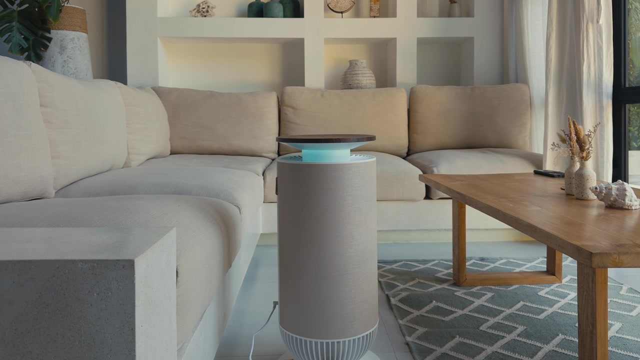 Air purifier operating with a soft blue light in the seating area of a modern bright living room with a beige sofa. A clean, stylish interior promoting healthy indoor air and comfortable living.