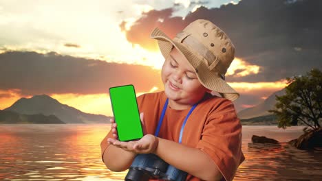 niño asiático con un sombrero y binoculares sonriendo y mostrando el teléfono inteligente de pantalla verde a la cámara. niño investigador examina algo en un lago, aventura de turismo de viaje, primer plano