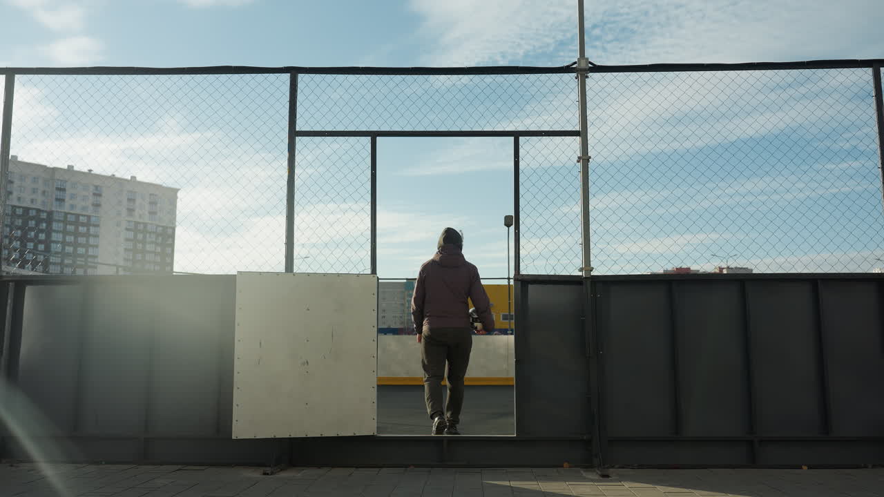 도시 스포츠 경기장에서 멀리 걸어가는 남자의 뒷면, 축구 공을 들고, 반이는 노란색 저장 컨테이너와 도시 건물을 배경으로