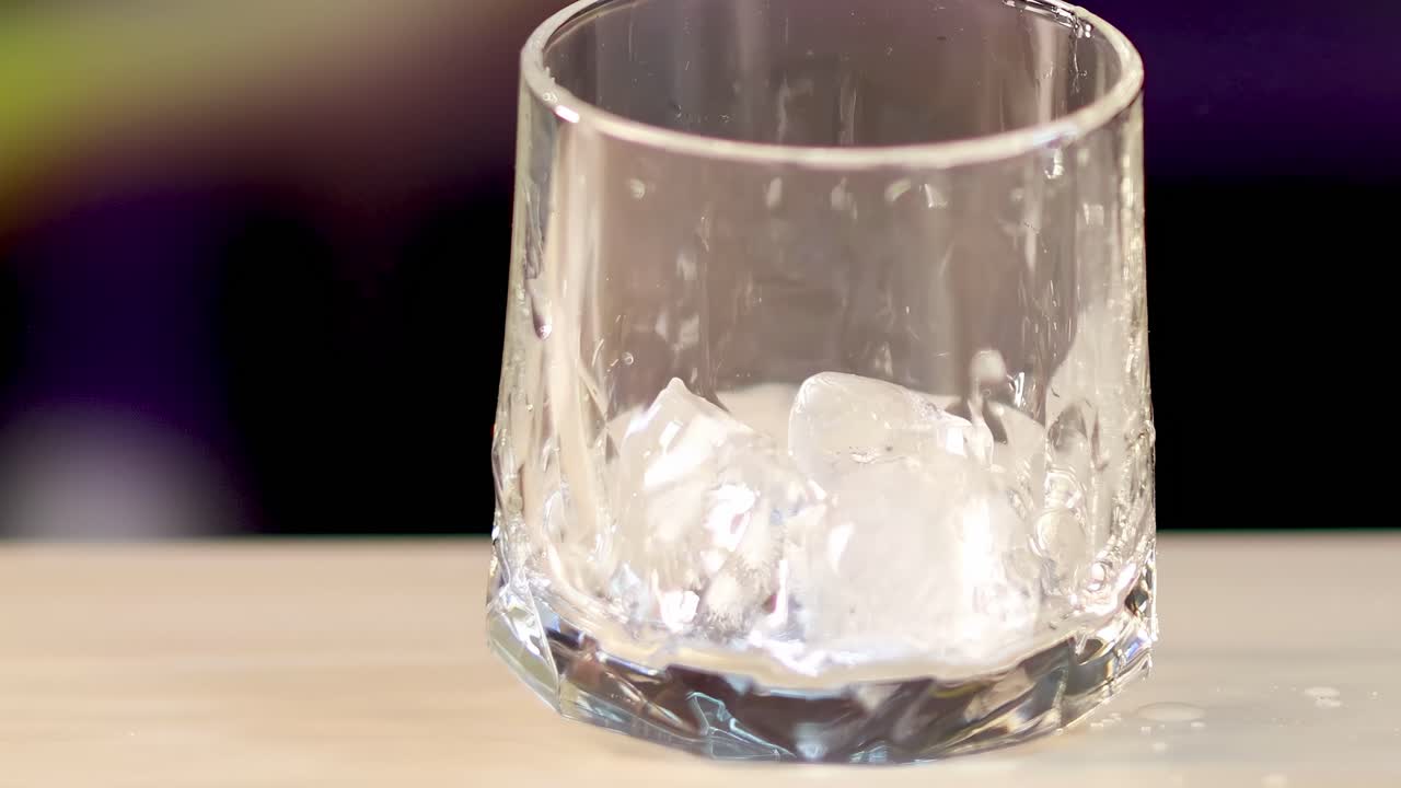 Ice cubes tumble and settle in a clear glass on a wooden table with a blurred background.