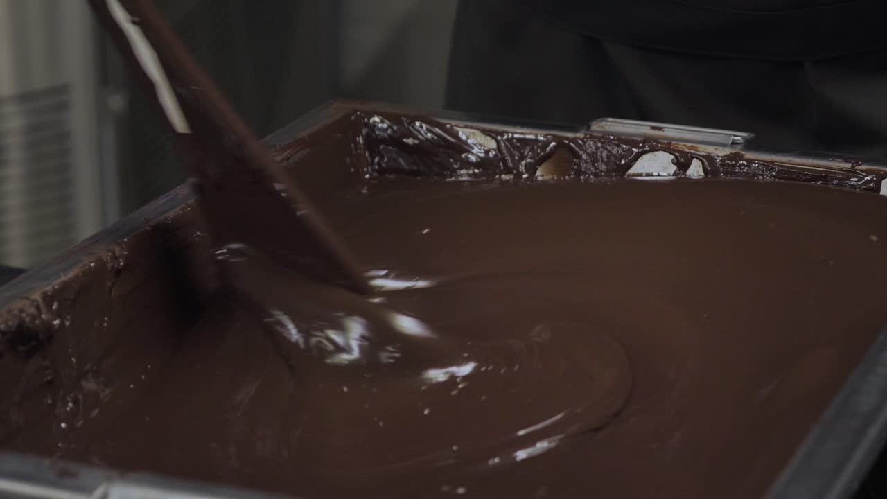 Close-up of thick melted dark chocolate pouring into metal tray creating glossy swirls in factory