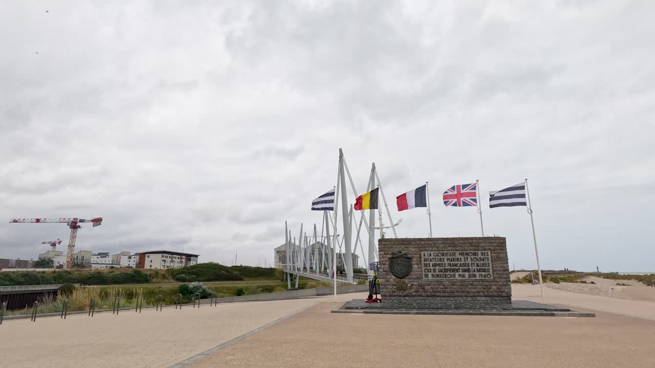 Wide shot of the Dunkirk Memorial with multiple flags waving in strong wind, overcast daylight, static camera, solemn and historical atmosphere