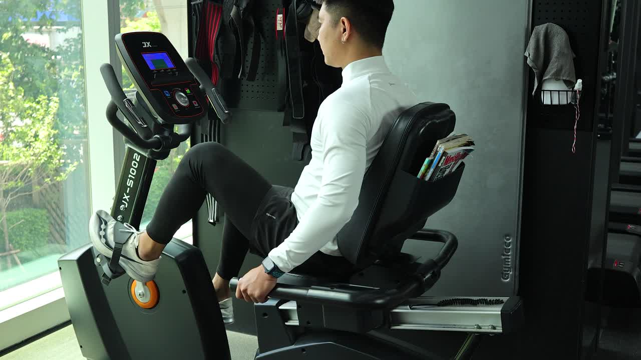 Asian man in athletic wear cycling on recumbent bike in bright, modern gym with daylight