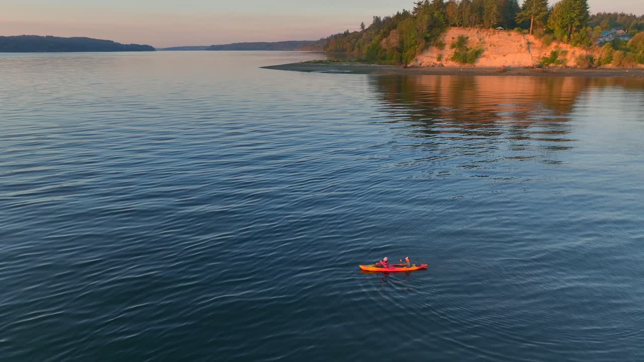 Aerial drone shot circling young couple sea kayaking paddling in bay to then reveal southworth ferry terminal pier near seattle washington at sunrise