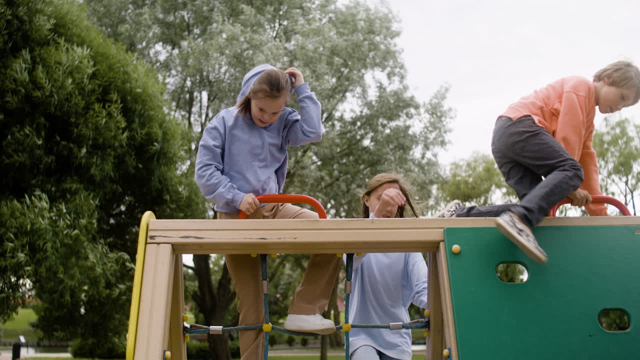 Little girl with down syndrome playing with her friends in the net of a children's park on a windy day. Then they sit on a wooden beam