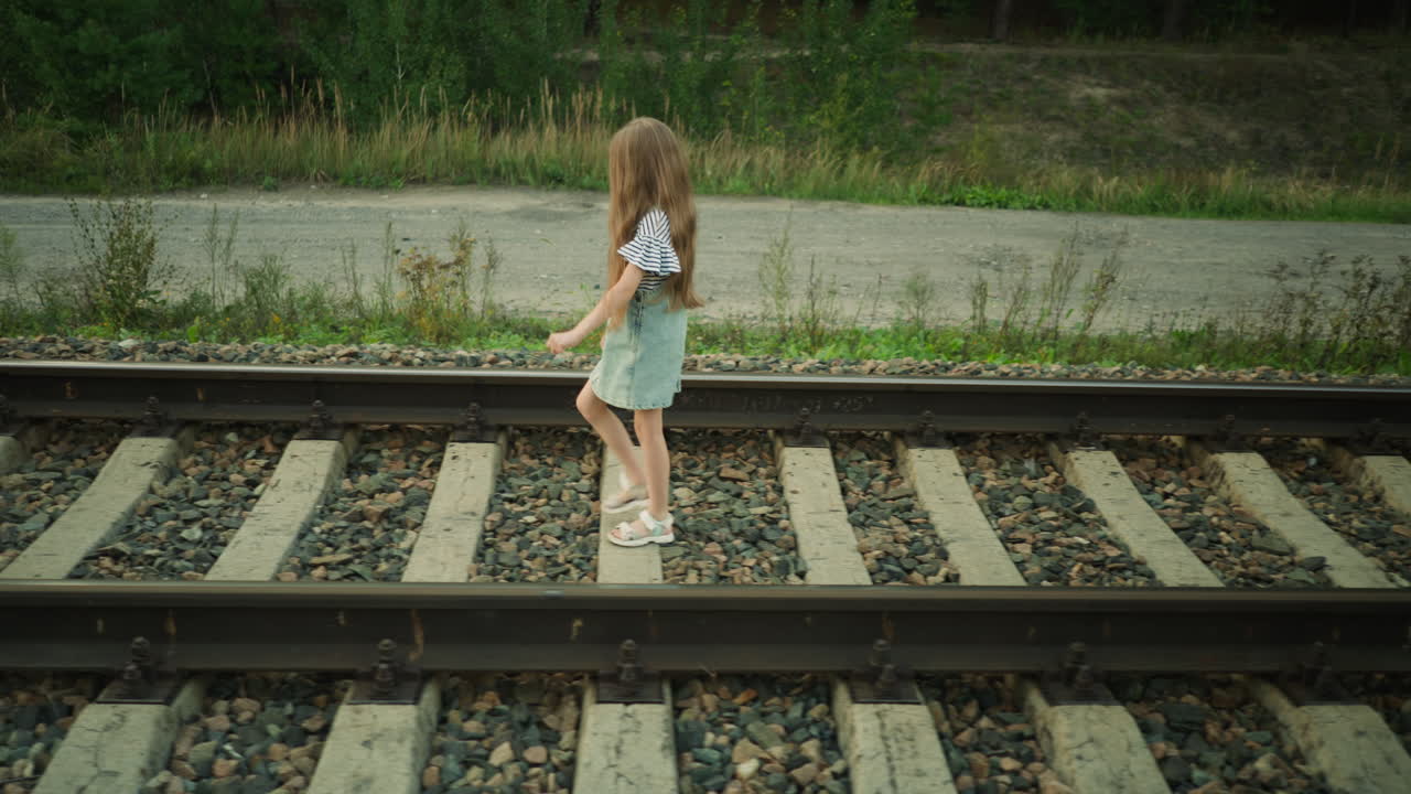 Side view of young girl walking confidently along rail track in rural environment, wearing striped shirt and denim dress, surrounded by green vegetation, gravel stones, and countryside road