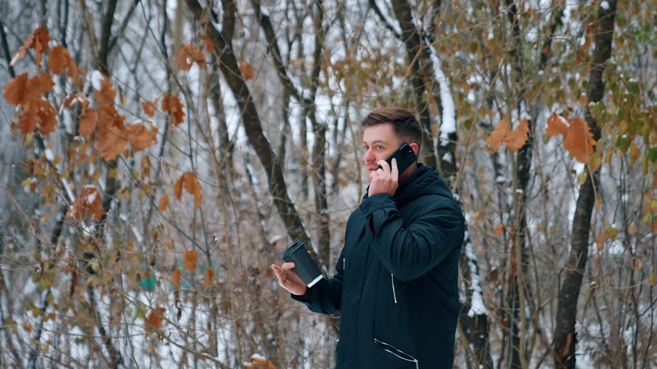 Handsome Caucasian male wearing black jacket speaks on the phone. Man with coffee cup in hand having a phone conversation outdoors in winter.