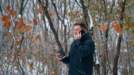 handsome caucasian male wearing black jacket speaks on the phone. man with coffee cup in hand having a phone conversation outdoors in winter.