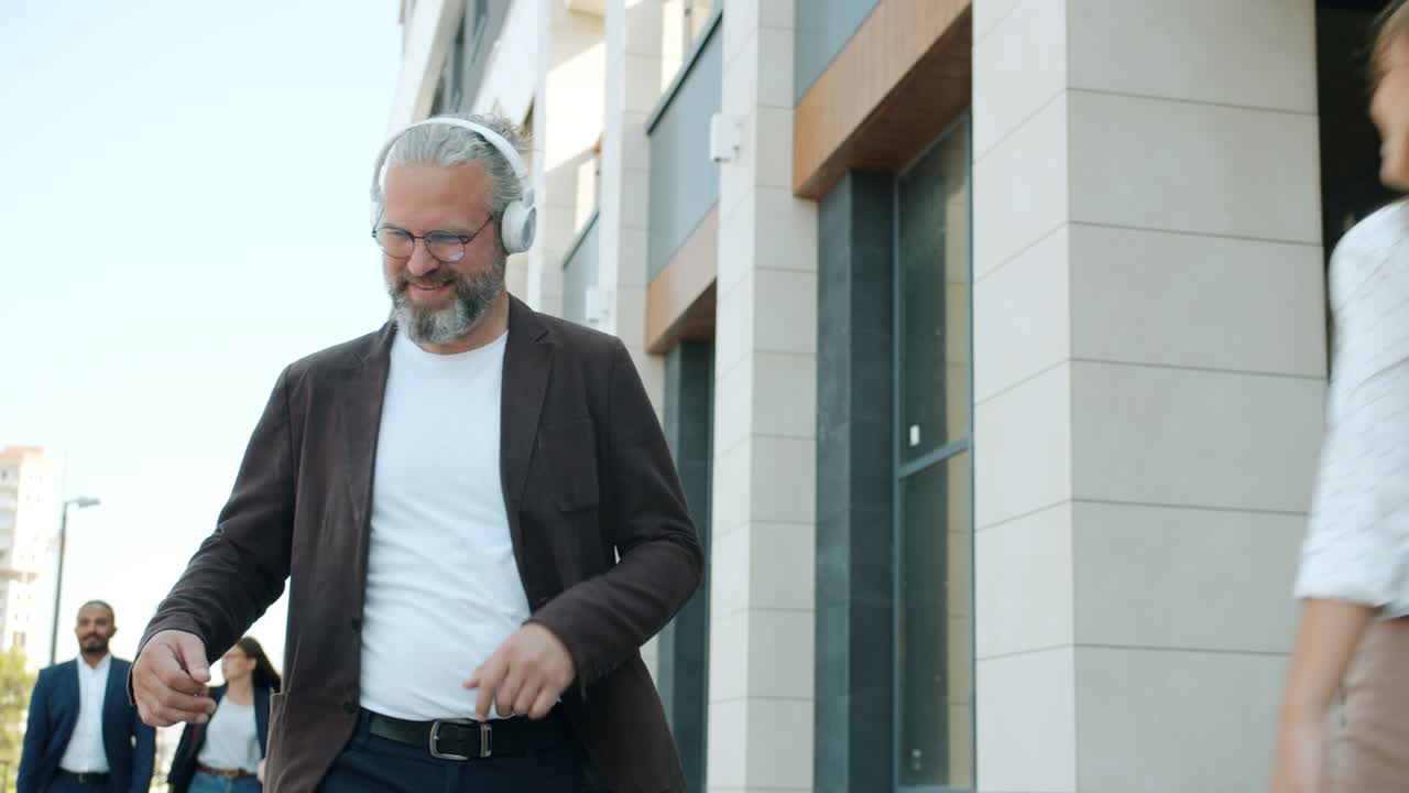 Careless mature man walking outdoors wearing headphones dancing enjoying music in urban street