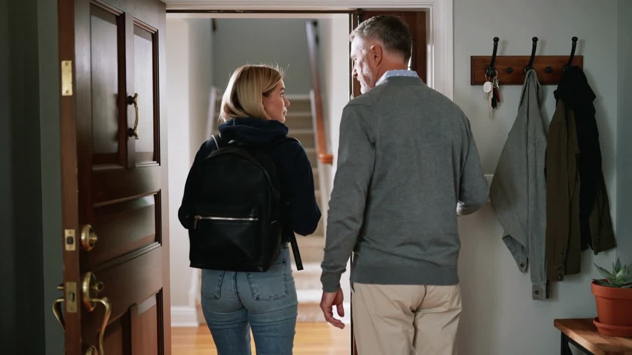 Apertura porta, donna con felpa, borsa nera e papà con maglione grigio che camminano verso le scale di casa, mentre parlano