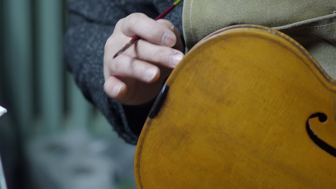 Close-up shot of luthier hand holding fine brush near the varnished back plate and ribs of a handcrafted violin, highlighting the wood grain, warm color tones, and artisanal craftsmanship in workshop