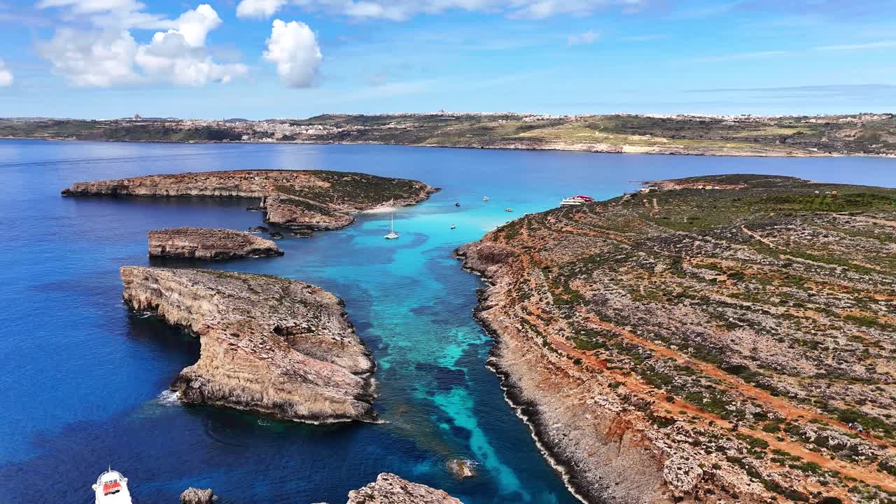 Luftfoto af klippekyst og lys turkis lagune ved Den Blå Lagune, Comino, Malta, med klart blåt hav, lagdelte klipper og små vige under solrig himmel.
