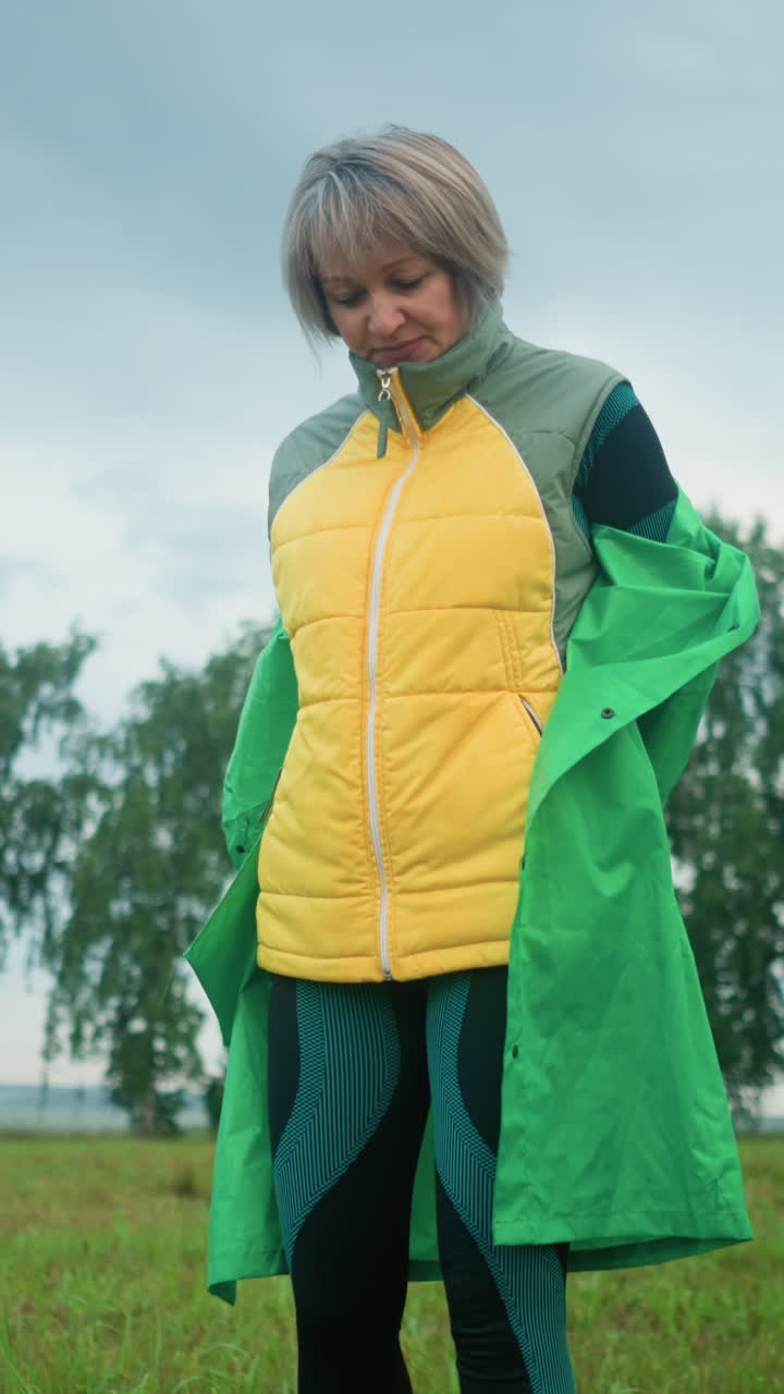 mulher de casaco amarelo sob um impermeável verde tira seu impermeável em um campo de grama nebulosa sob um céu nublado com árvores à distância
