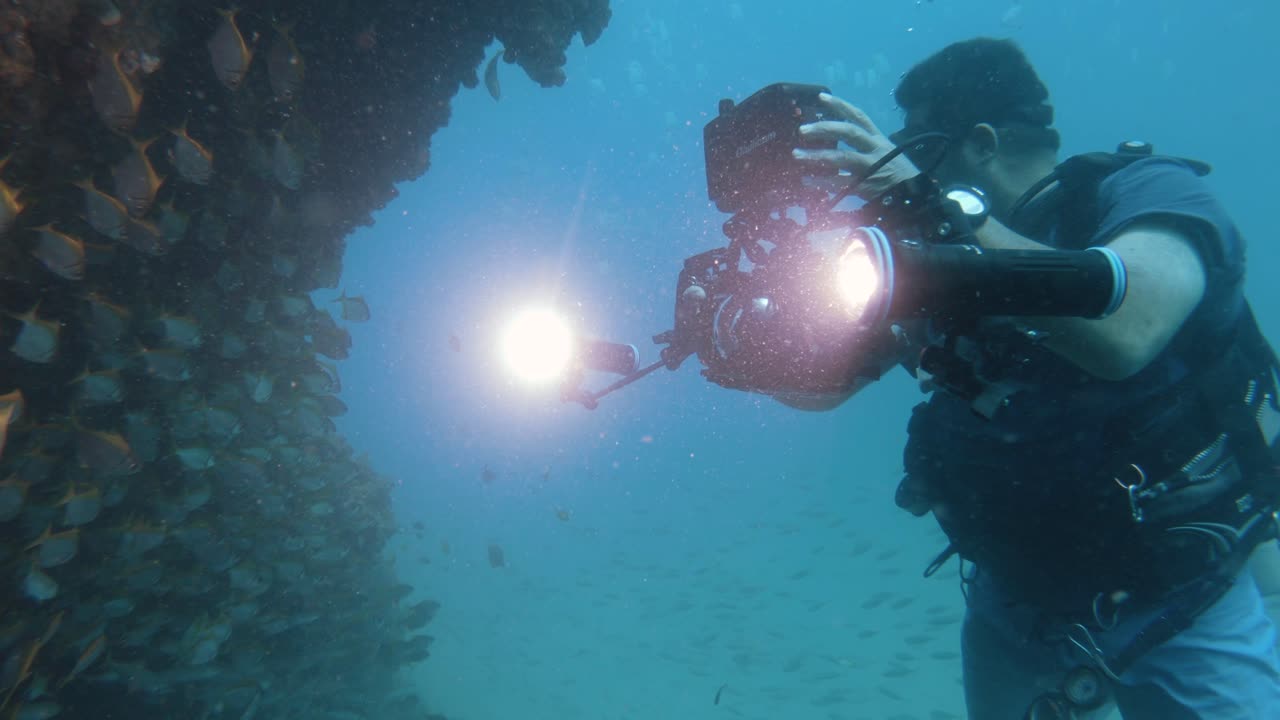 A scuba diver operating an underwater camera set-up attached with bright lights swims slowly through a large school of darting fish as he captures and records the underwater action