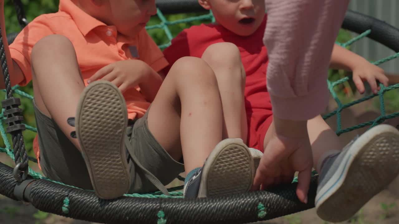Bambini che si divertono ad oscillare allegramente all'aperto, giovani seduti su altalene circolari alla luce del sole, bambini felici seduti tranquillamente su altalene rotonde mentre un adulto le modifica e le spinge delicatamente alla luce del giorno