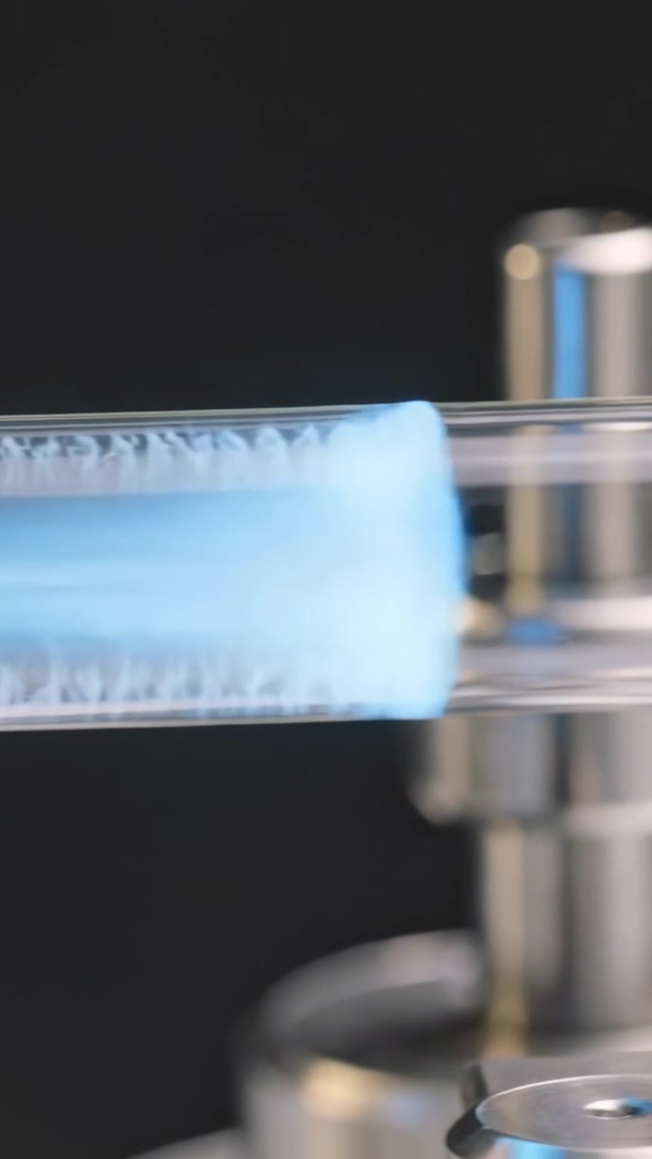 Video verticale: Plasma blu che si accende a sinistra e si muove verso destra lungo un tubo trasparente su un banco, con un supporto a morsetto.