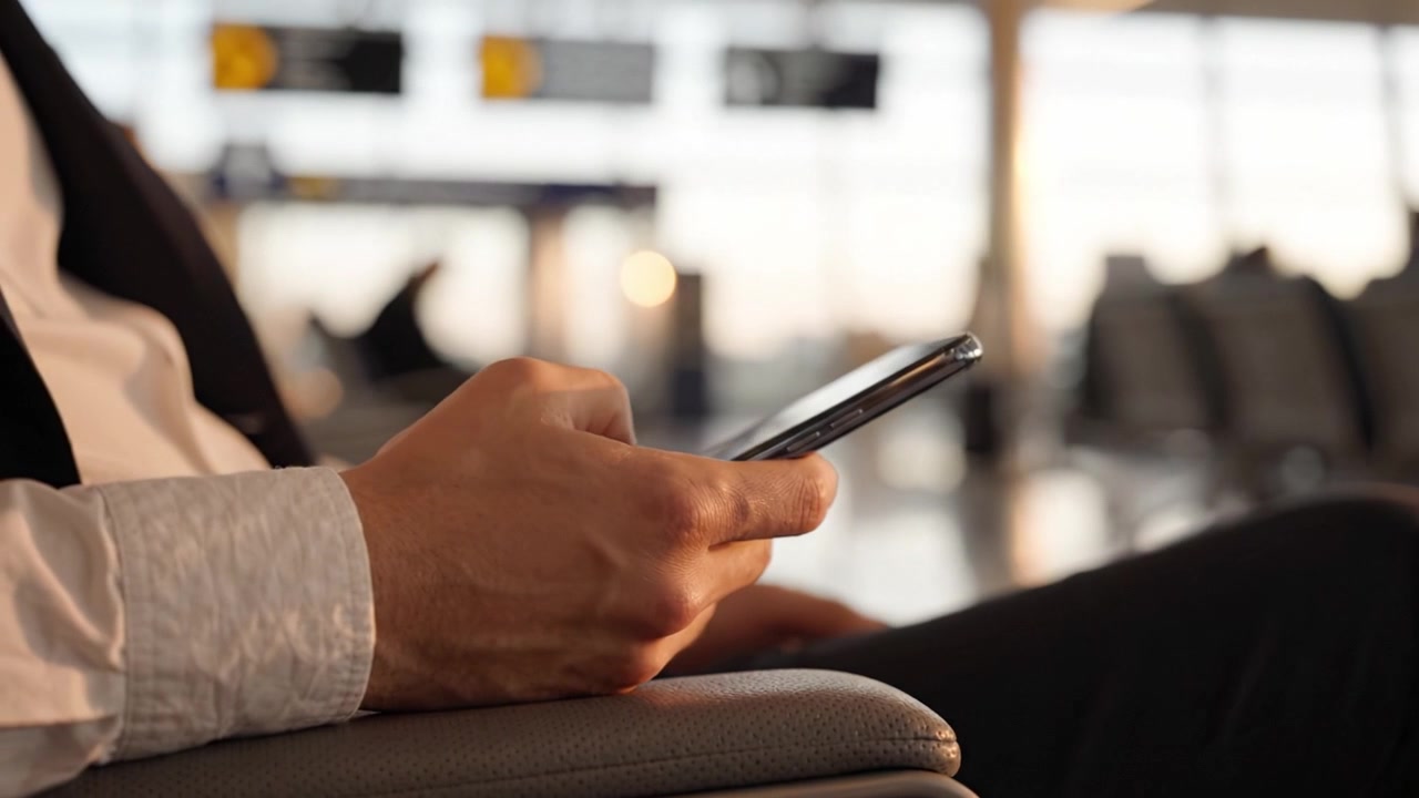 A Person Sitting in an Airport Lounge, Engrossed in Their Smartphone, Capturing Moments of Travel Excitement and Anticipation Amidst a Busy Terminal