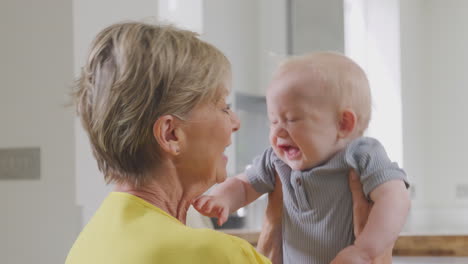 Grandmother-Looking-After-And-Playing-With-Laughing-Baby-Grandson-Sitting-At-Kitchen-Table