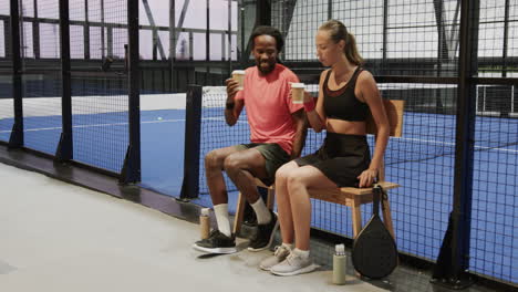 diverse athletes enjoying coffee break after padel tennis match, sitting on bench, on indoor court