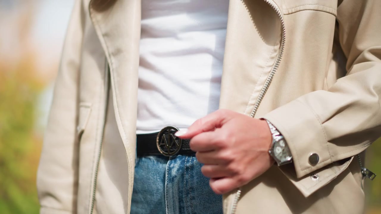 Primo piano di una donna che indossa una giacca beige, una camicia bianca e jeans blu, con la mano che aggiusta la cerniera. Un orologio al polso è visibile, insieme a dettagli come i motivi delle cuciture nel tessuto.