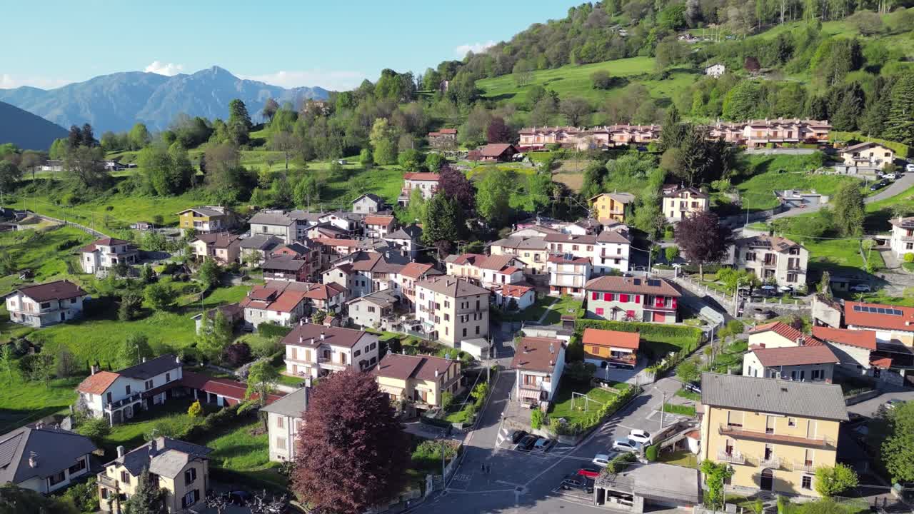 A drone slowly flies backward over San Fedele near the city of Como, revealing traditional rooftops, narrow streets, green hills, and towering mountains in the summer sun