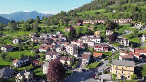 a drone slowly flies backward over san fedele near the city of como, revealing traditional rooftops, narrow streets, green hills, and towering mountains in the summer sun
