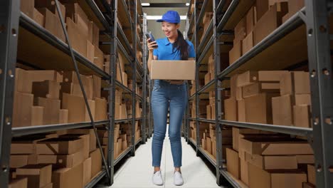 full body of asian female courier in blue uniform using smartphone while delivering a carton in warehouse