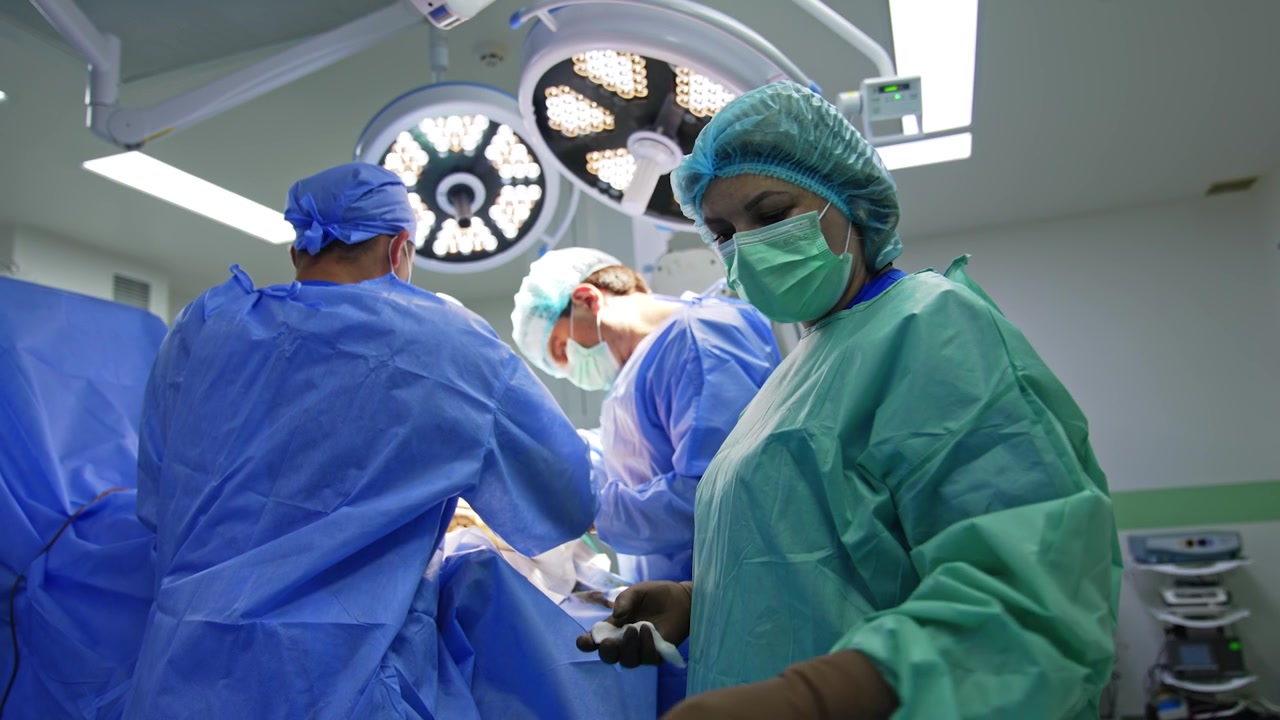 Un'infermiera strumentista donna, che indossa uniforme protettiva e mascherina, passa gli strumenti ai chirurghi principali. Un intervento di chirurgia traumatologica procede in una moderna sala operatoria.