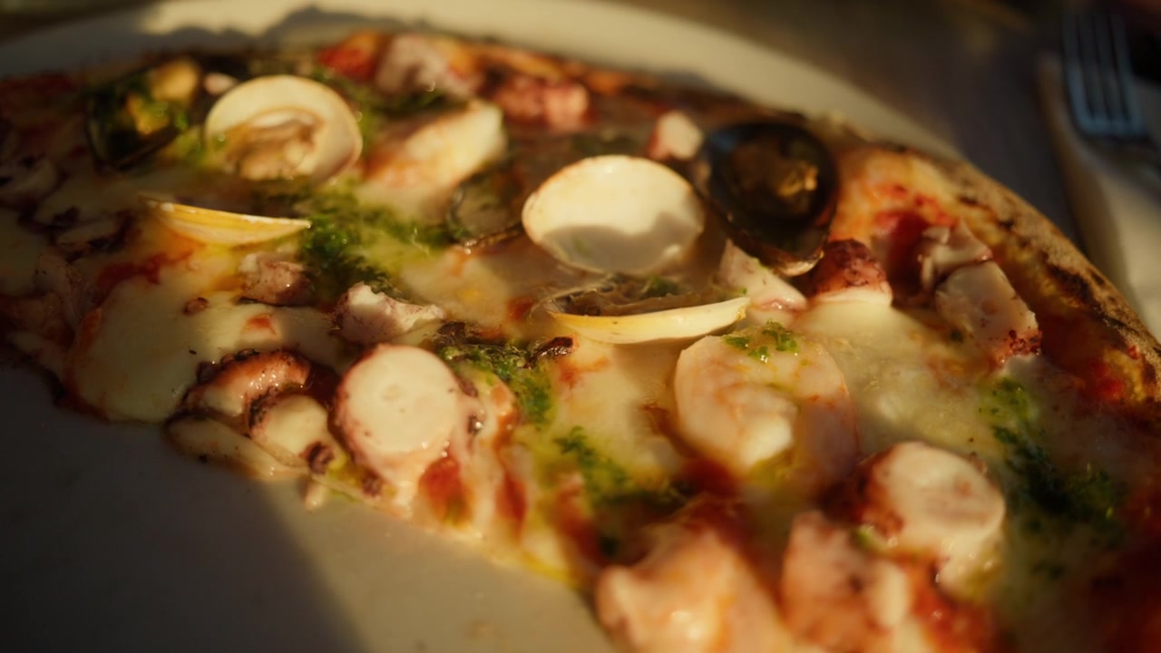 Primo piano di una fetta di pizza ai frutti di mare su un piatto, con gamberi, cozze, vongole e polpo, illuminata dalla luce del tramonto.