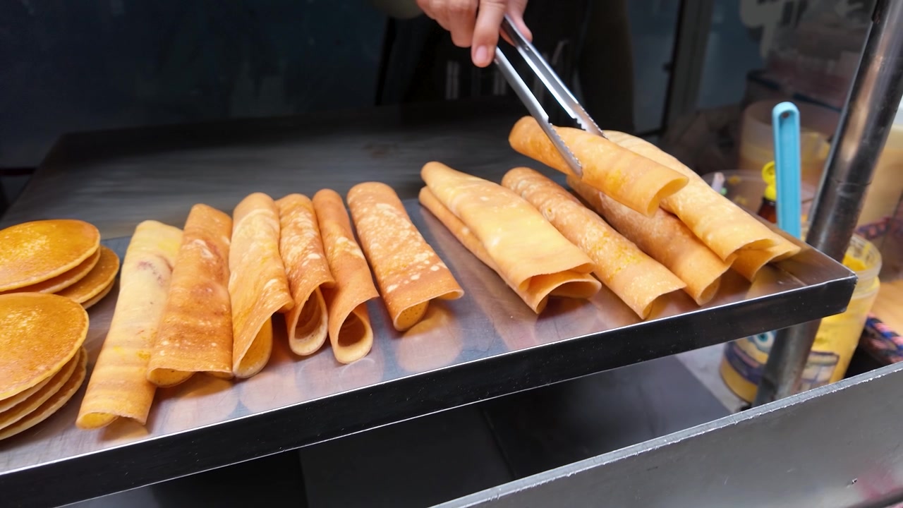 Bidikan close-up seorang penjual menggunakan penjepit untuk menata crepes emas di wajan logam.