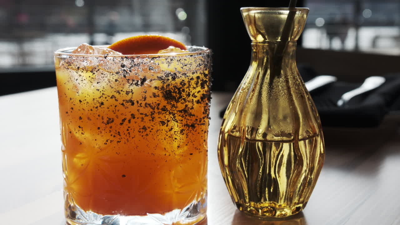 Beautifully garnished orange cocktail with a spiced rim sits on a table next to a unique golden carafe