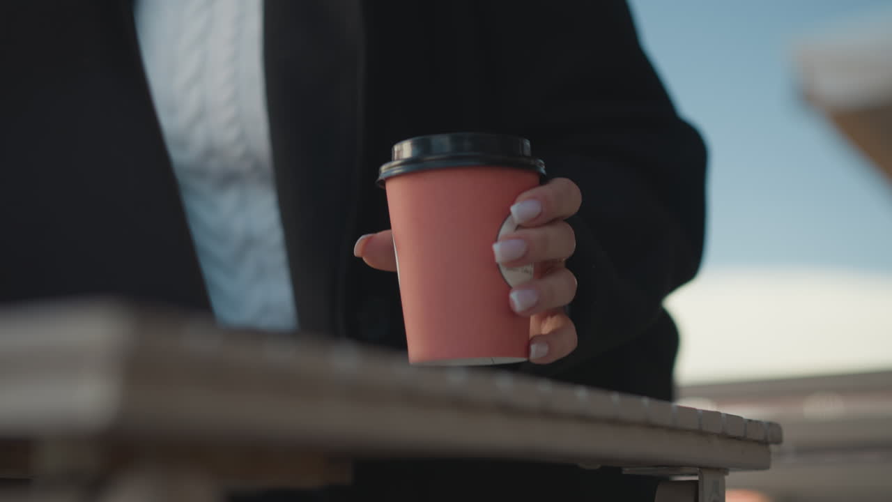 노트북 앞 테이블 위에 커피 컵을 놓고 은 손을 가진 누군가의 클로즈업, 불분명한 구조의 흐릿한 배경, 야외 테이블에 앉으려는 사람