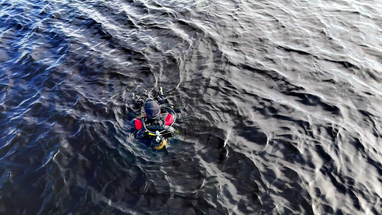 Subacqueo con attrezzatura completa e dettagli rossi sulle spalle galleggia tranquillamente sulla superficie di acqua scura increspata, circondato da onde che riflettono mutevoli giochi di luce