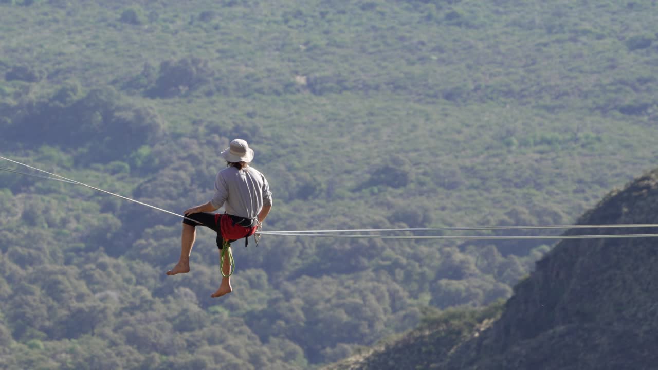 A man contemplates the forest below while sitting on a highline. Beauty in nature, extreme sport, 4k, slow-motion