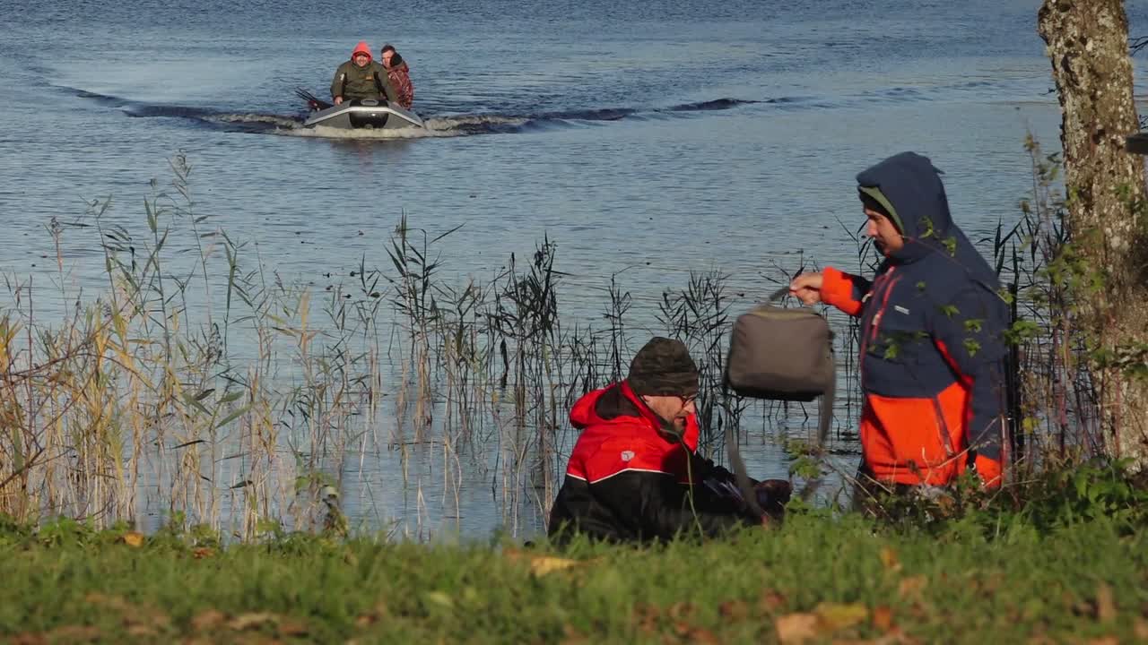 Fishermen in a boat on a blue lake, casting lines and catching fish on a calm day. The water reflects the autumn landscape, enhancing the serene and seasonal feel.