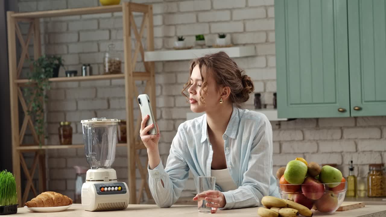 Woman in kitchen looking at her phone and drinking water