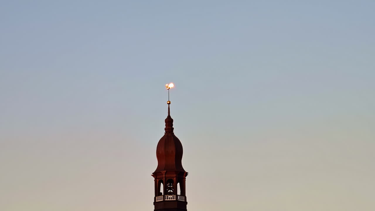 Historic church tower in Riga silhouetted at dusk with a glowing weather vane against a calm sky