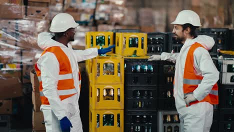 A-man-with-Black-skin-in-a-white-protective-uniform-in-an-orange-vest-communicates-with-his-male-colleague-with-a-beard-while-they-are-working-and-being-in-a-large-recycling-plant-for-plastic-and-glass-bottles