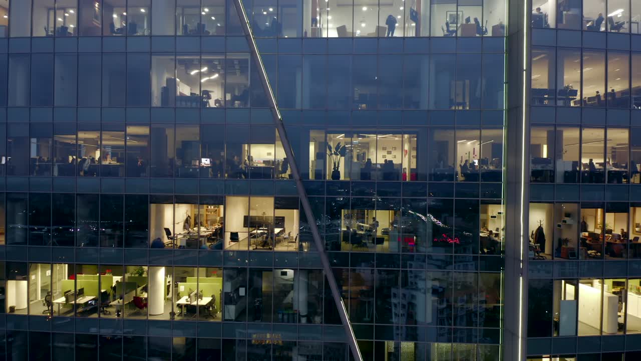 una toma cinematográfica de un dron en un rascacielos nocturno con ventanas de oficinas y gente de negocios trabajando hasta tarde en las computadoras.