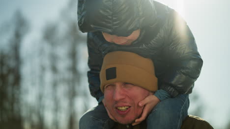 close up of a father carrying his child on his shoulders, as the child play with his chin and he looks tired as his face becomes red because of the child we